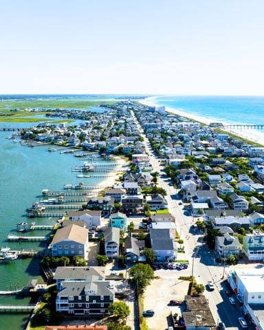 Aerial view of coastal barrier island with residential homes, docks, beach, and pier extending into turquoise water