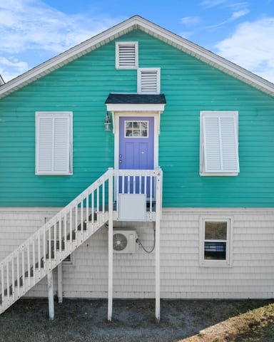 Turquoise cottage with white trim, purple front door, and white railed deck stairs, neighboring beach houses visible