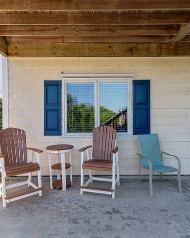 Covered patio with Adirondack chairs, small table, and blue entry door on cream-colored house with exposed beam ceiling
