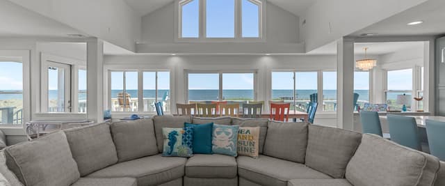 Bright open-plan living room with curved gray sectional sofa, vaulted ceiling, and expansive ocean views through multiple windows and glass doors