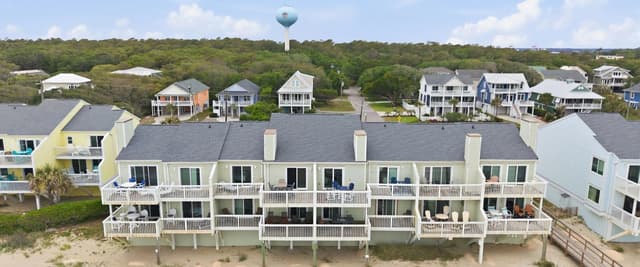 Beachfront row of elevated vacation rental homes with white railings and decks, sandy beach in foreground, water tower and tree-covered landscape beyond.