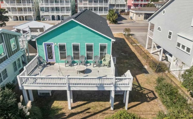 Turquoise beach cottage with white deck and Adirondack chairs, surrounded by other vacation homes in a coastal neighborhood.