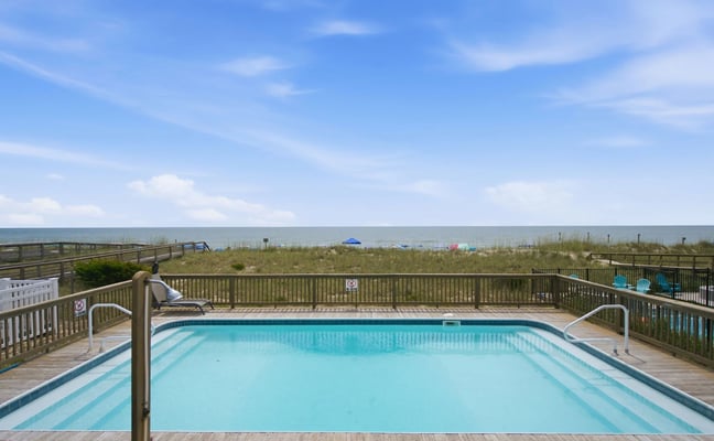 Elevated pool deck with light blue swimming pool overlooking beach, dunes, and ocean under clear sky