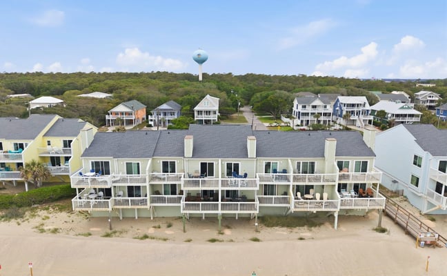 Beachfront row of elevated vacation rental homes with white railings and decks, sandy beach in foreground, forested residential area behind.