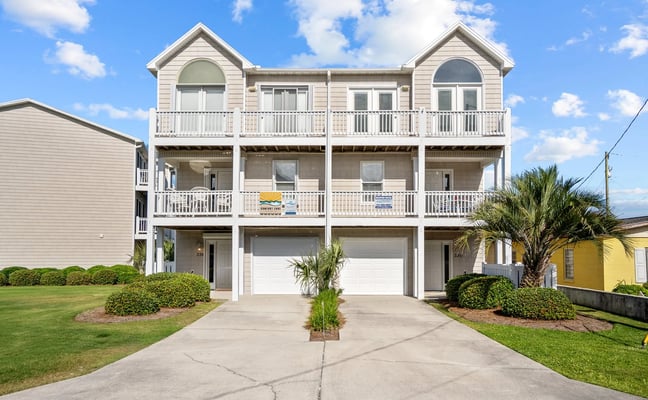 Beige three-story beach house with white railings, dual garages, and palm trees in front yard
