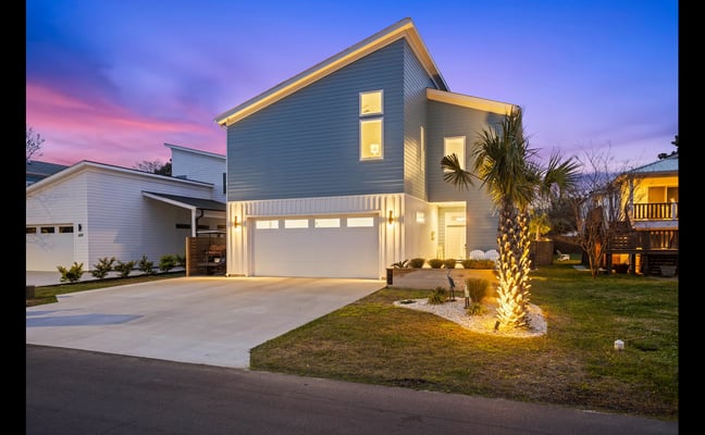 Modern gray house with illuminated garage and windows at dusk, palm tree in front yard, neighboring homes visible.