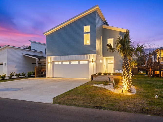 Modern gray house with illuminated garage and windows at dusk, palm tree in front yard, neighboring homes visible.