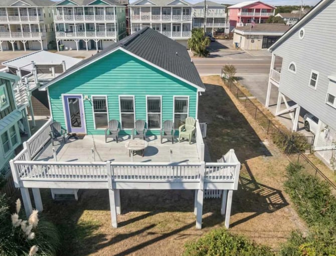 Turquoise beach cottage with white deck and Adirondack chairs, surrounded by other vacation homes on a residential street.